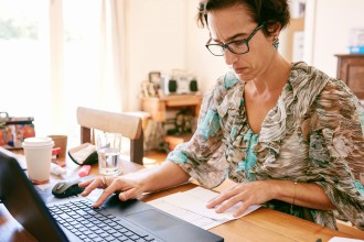 Older business woman busy entering digits on her new laptop computer from a printed list that she's reading while wearing glasses