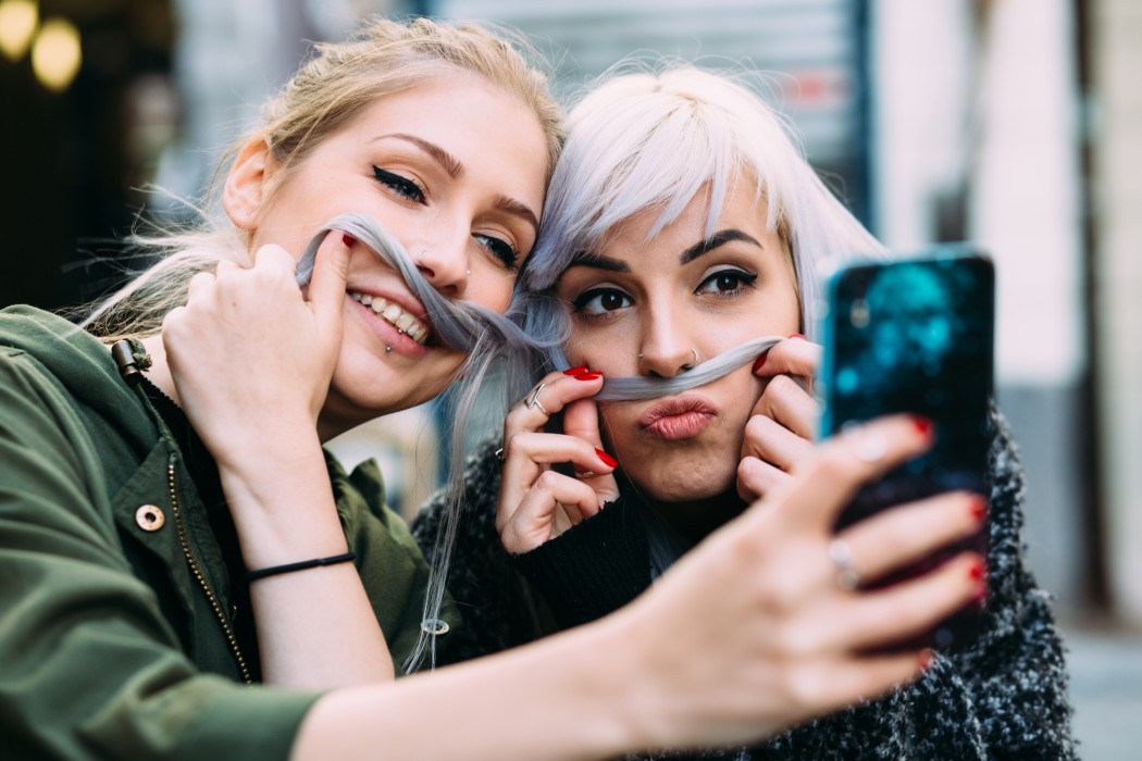Two young women taking a selfie together and using one of their hair to make a moustache.
